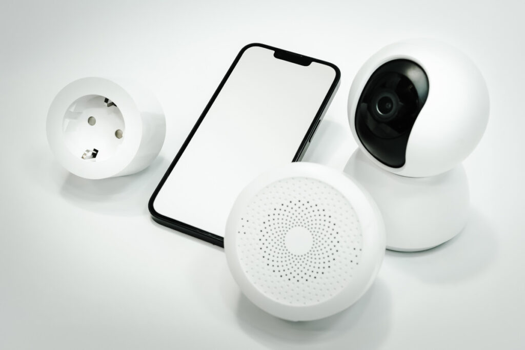 Dispozitive inteligente pentru securitate: camera, telefon si difuzor, alaturi de un detector de fum wireless, pe fundal alb.